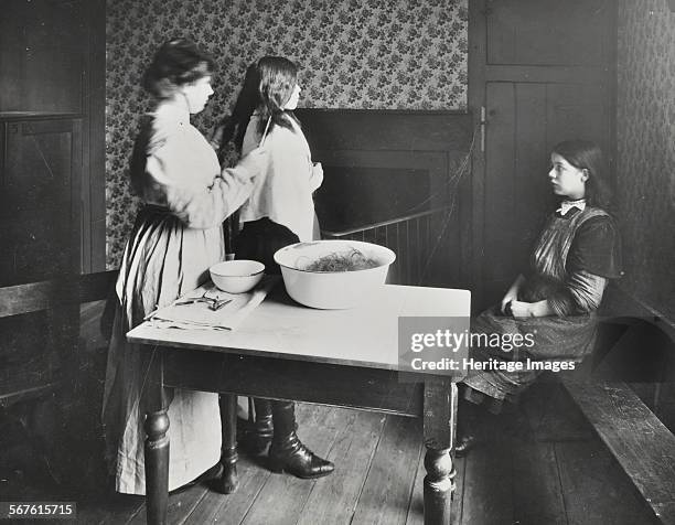 Nurse examines girls' hair, Central Street Cleansing Station, London, 1914. Woman checking for head lice; on the table is a bowl of cut hair. Most...