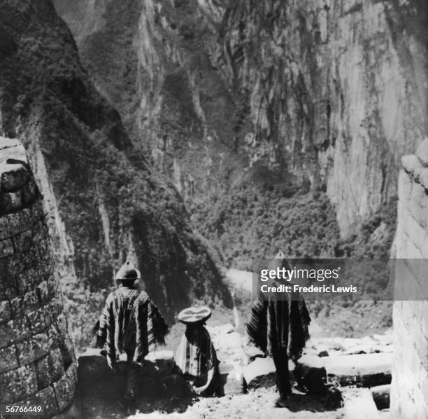 Three Peruvian Indians stand in traditional dress on a masonry ledge of the ancient Incan city of Machu Picchu and look out over a deep valley at one...