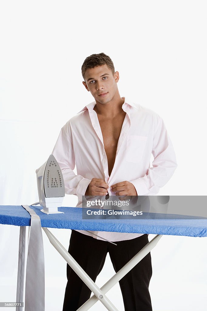 Portrait of a young man standing in front of an ironing board and buttoning his shirt