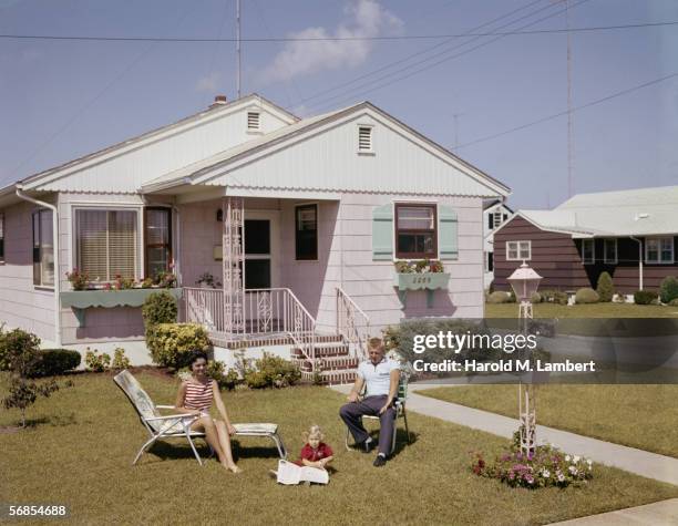 The Storr's and their young daughter sitting in the front garden ona sunny day, 1960. The girl is looking through a newspaper.