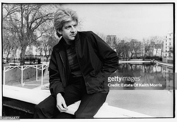 Outdoor portrait of musician Nick Lowe sitting on the edge of a bridge overlooking a river, circa 1990.