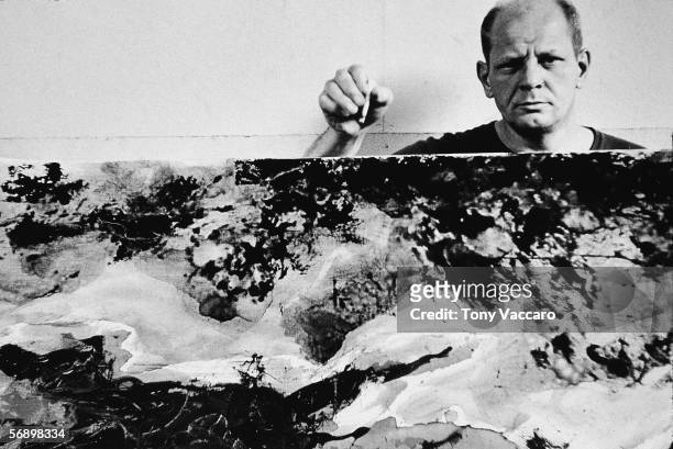 American abstract expressionist painter Jackson Pollock holds a cigarette above and behind one of his paintings in his studio at 'The Springs,' East...