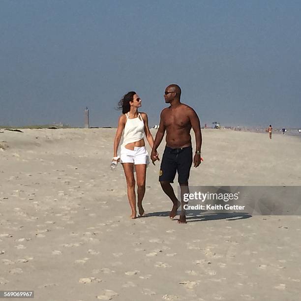 Man and woman couple holding hands and looking each other in the eye whole strolling on a beautiful sand beach against an expansive blue gray sky -...