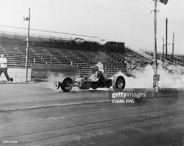 Unidientified drag racer drives a three-wheeled dragster off the starting line at a race track as two men look on, mid 20th Century.