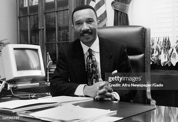 Hands folded on the table, Kweisi Mfume sitting in an office, Japan, 1990. There is a computer monitor on the desk.