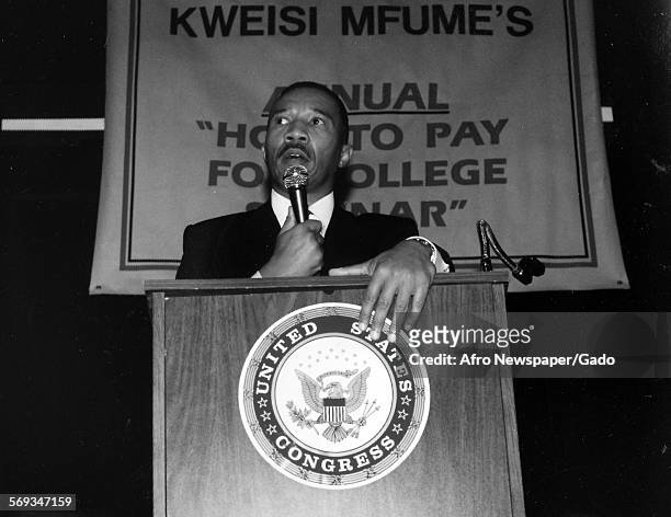 Kweisi Mfume holding a microphone and standing at a podium with a United States Congress seal, 1991.