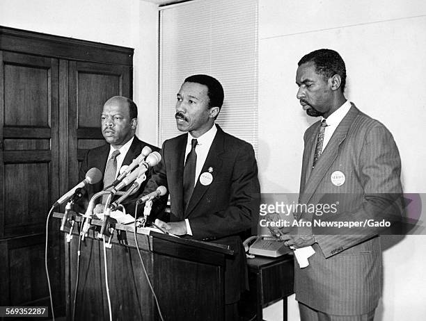 Standing in between two other men, Kweisi Mfume delivering a speech at the podium, 1990.