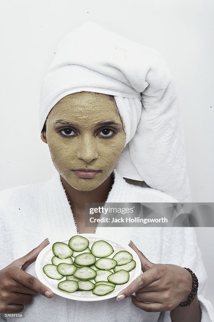 Portrait of a young woman with a facial mask holding a plate of sliced cucumber