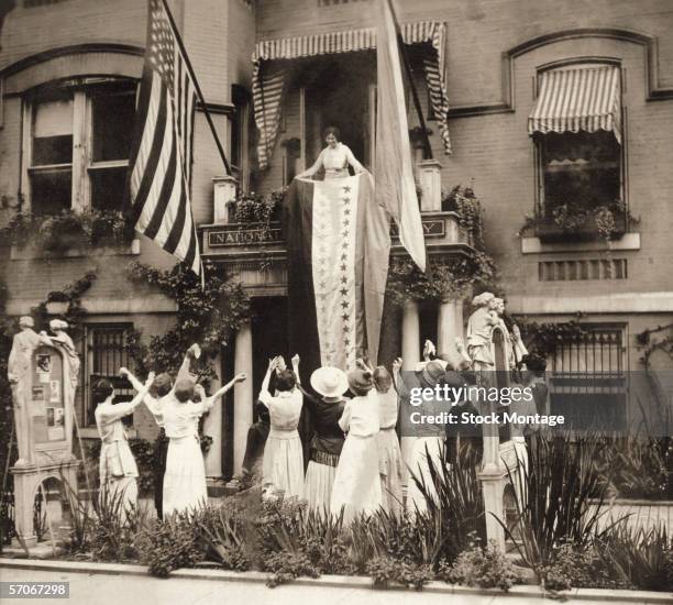 As women stand and cheer, American suffragist Alice Paul stands on a balcony at the National Women's Party headquarters and unfurls a banner in...