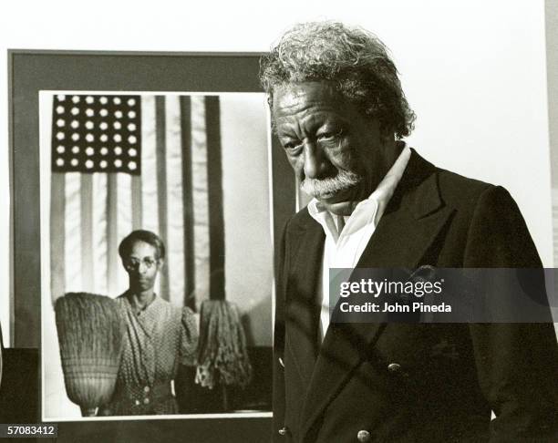 American photographer and film director Gordon Parks stands next to one of his most famous images, 'American Gothic,' which depicts a cleaning woman...