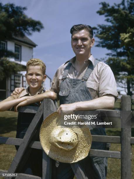 Father and son leaning on a gatepost on a farm, 1947. The father is wearing dungarees and has his arm around the boy.