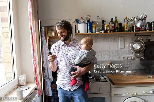 father holding baby son while using smartphone. - two parents photos et images de collection