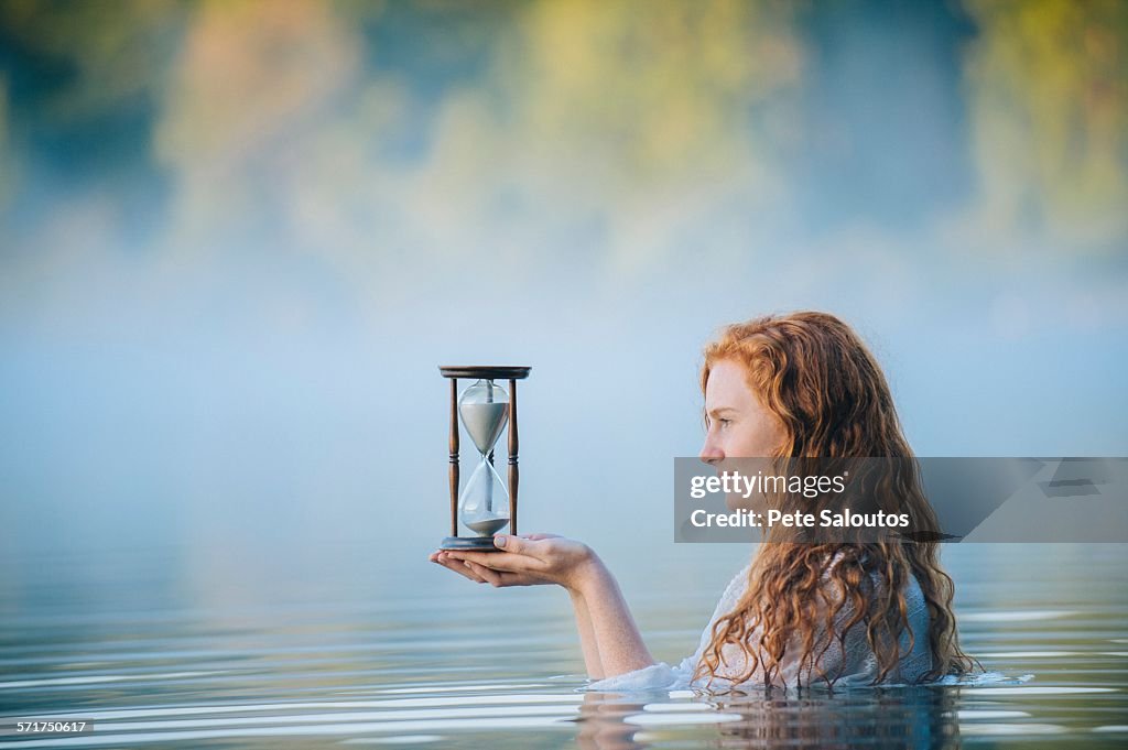 Young woman standing in misty lake gazing at hourglass