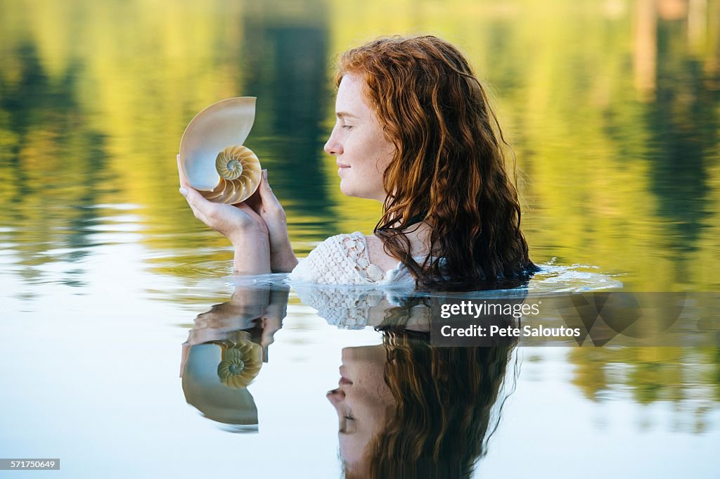 Head and shoulders of young woman with long red hair in lake gazing at seashell
