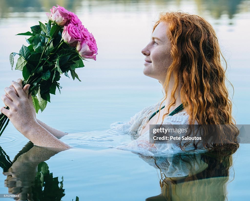 Head and shoulders of young woman with long red hair in lake gazing at bunch of pink roses