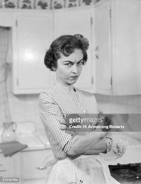 1950s: Housewife in kitchen, arms folded, with serious expression.