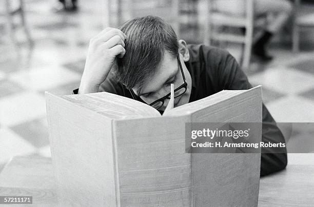 1950s: Man sitting at library table, scratching head, with large book open in front of him.