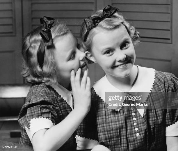 girl (12-13) indoors whispering into ear of twin sister, (b&w) - 20th century stock pictures, royalty-free photos & images