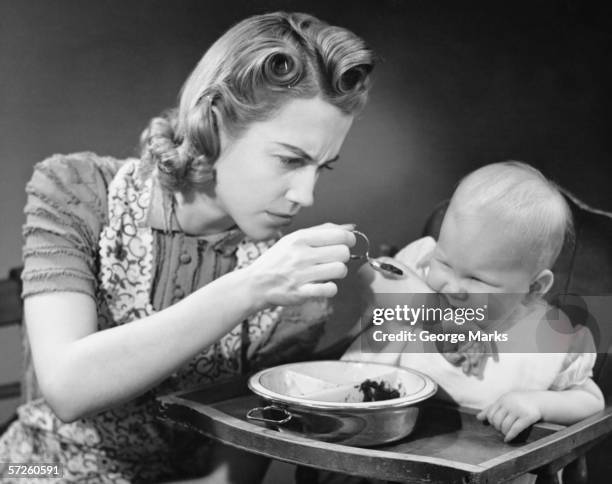 mother trying to feed unwilling baby (9-12 months), (b&w) - housewife stock pictures, royalty-free photos & images