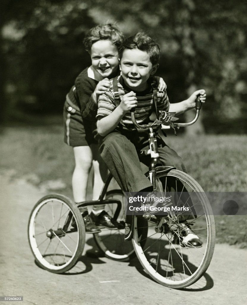 Two young boys (6-7) riding tricycle in park, (B&W), portrait