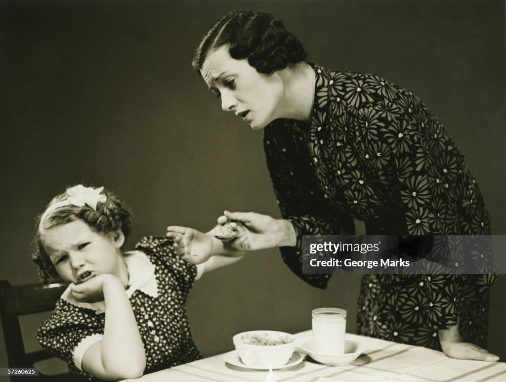 Mother trying to make daughter (4-5) finish food at table, (B&W)