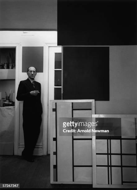 Portrait of Dutch modernist painter Piet Mondrian as he stands and smokes a cigarette near some of his paintings, New York, January 17, 1942.