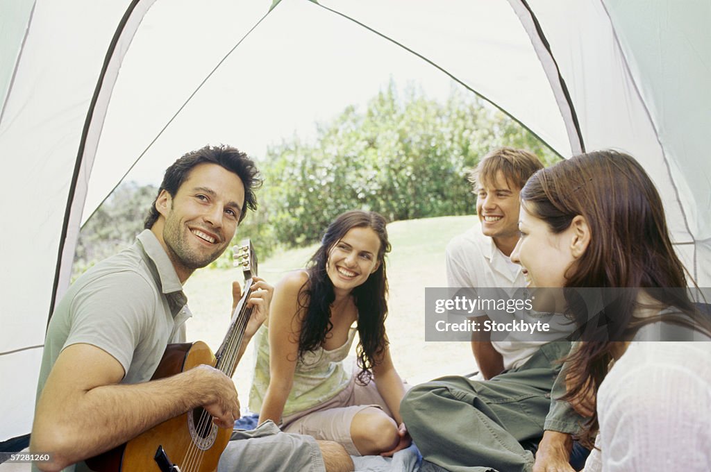 Young man sitting with his friends and playing guitar inside a tent