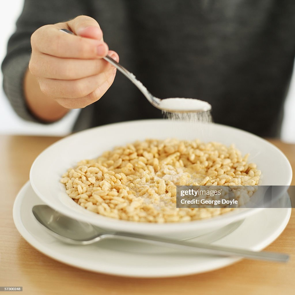 Person sprinkling sugar over a bowl of rice crispy