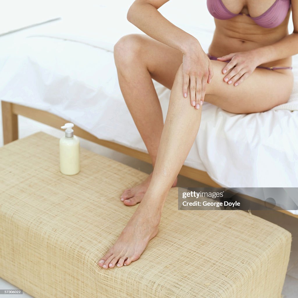 Close-up of a woman applying lotion on her leg sitting on a bed in underwear