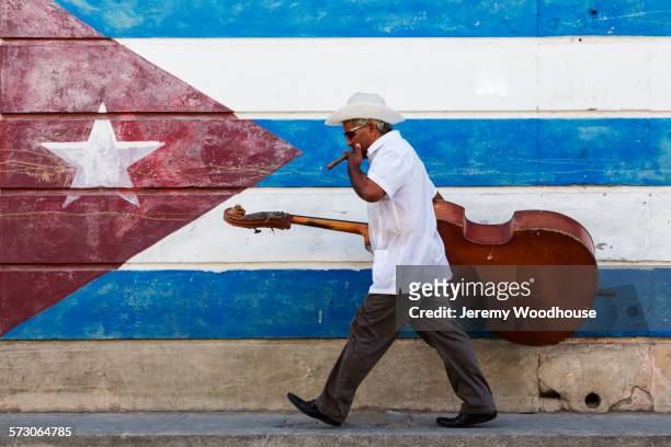 hispanic musician carrying upright bass in front of cuban flag mural - caribbean musical instrument stock pictures, royalty-free photos & images