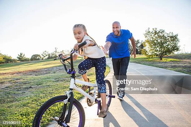 father teaching daughter to ride bicycle in park - adult riding bike through park stock pictures, royalty-free photos & images