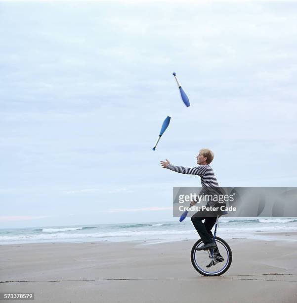 man juggling pins on unicycle on beach - giocoliere foto e immagini stock