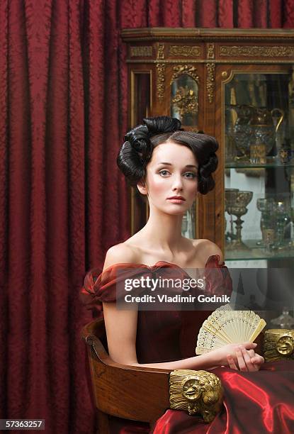 elegant young woman seated in red ball gown and holding fan - woman in evening dress stock-fotos und bilder