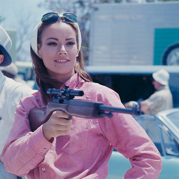 French actress Claudine Auger pictured in character as Domino Derval, wearing a pink shirt and holding a rifle during filming of the James Bond film...