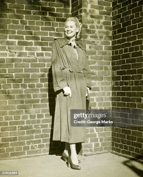 woman wearing mackintosh standing at brick wall outdoors, (b&w) - vintage rainwear stockfoto's en -beelden