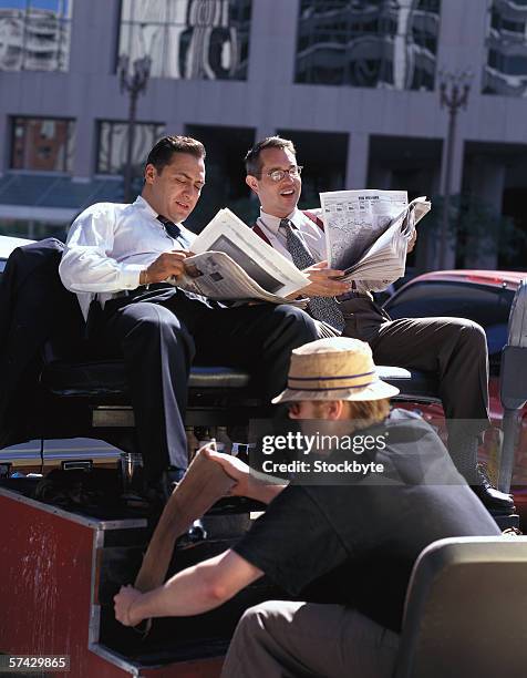 two young businessmen getting their shoes polished while reading the newspapers outdoors - 靴をみがく ストックフォトと画像