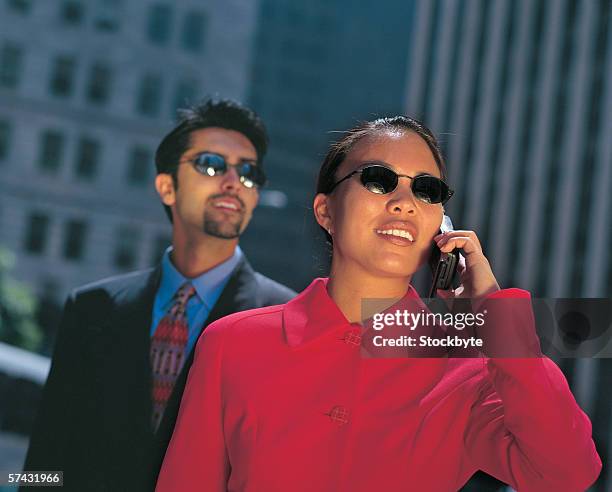 close up of a woman wearing sunglasses talking on a mobile phone with a man standing behind - business woman in red suit jacket stock pictures, royalty-free photos & images