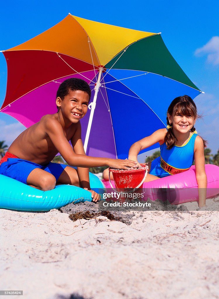 Two young children sitting on flotation tubes playing with sand on the beach