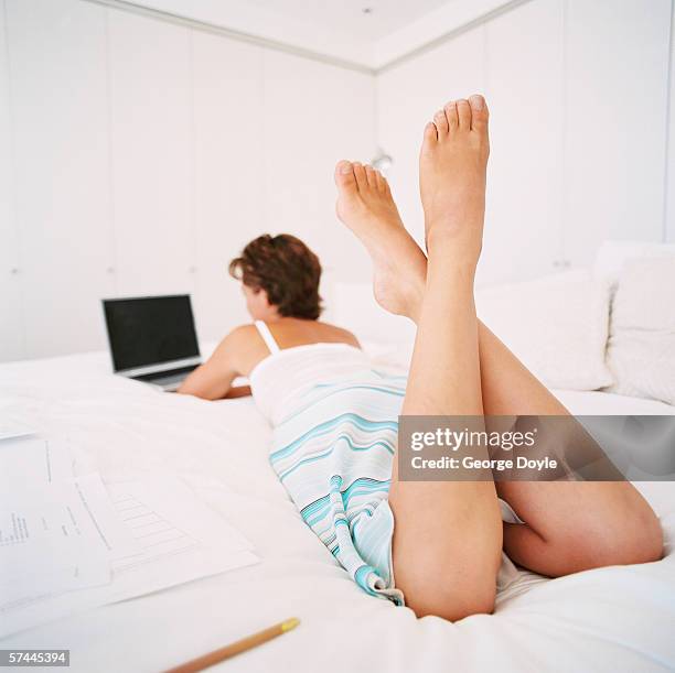 view from behind of a woman lying on her stomach on a bed and working on a laptop computer - woman lying on stomach with feet up foto e immagini stock