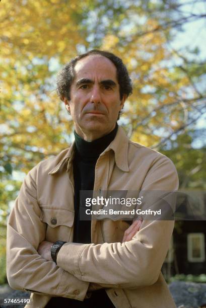 Portrait of American novelist Philip Roth, dressed in a turtleneck and jacket, who stands outdoors with his arms crossed, probably 1980s.