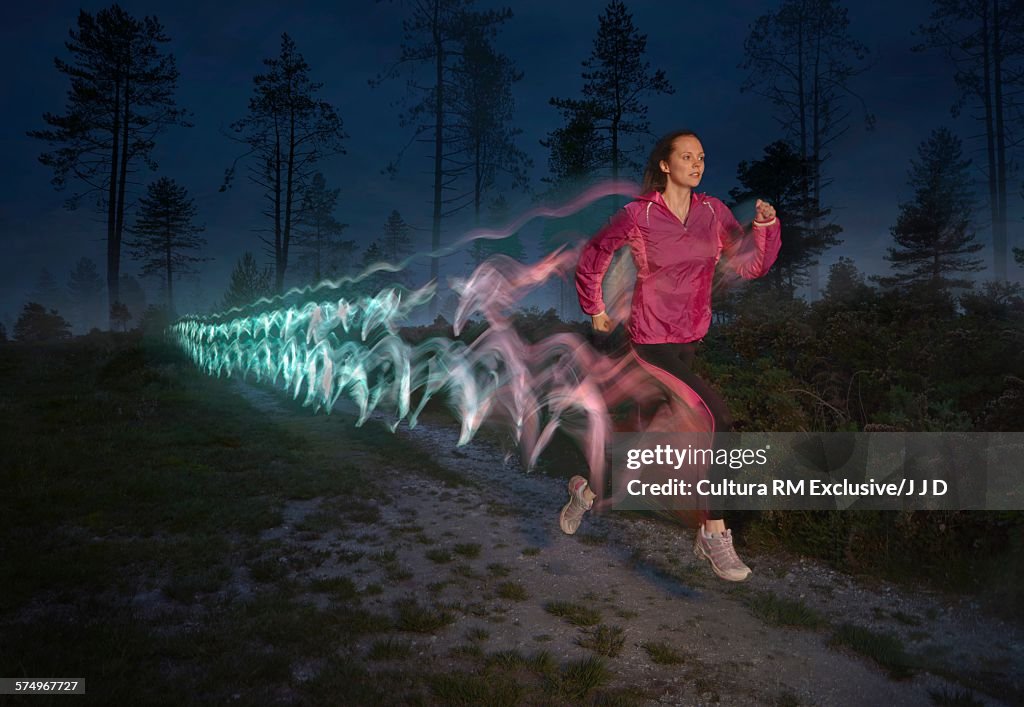 Young woman followed by light trails running on forest dirt track at night