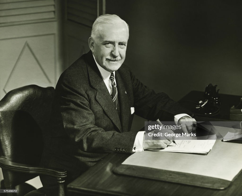 Senior businessman signing documents at desk, (B&W), portrait