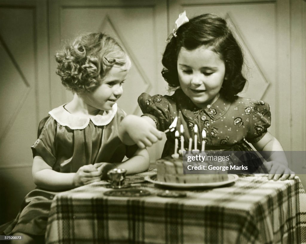 Two girls (3-4), (4-5) sitting at small table with birthday cake, (B&W)