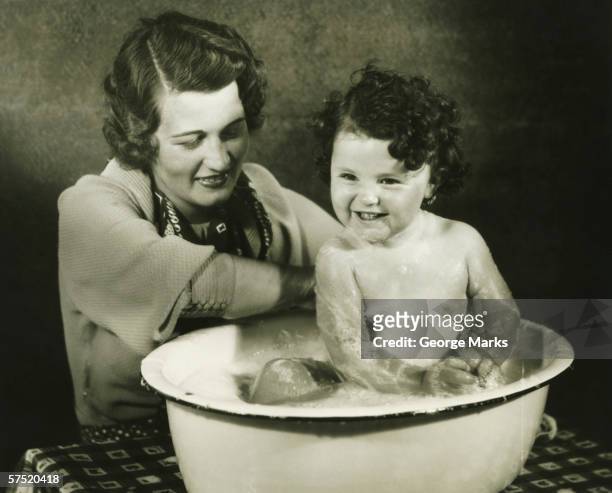 mother bathing daughter (12-18 months) in basin, (b&w) - 1930 stockfoto's en -beelden