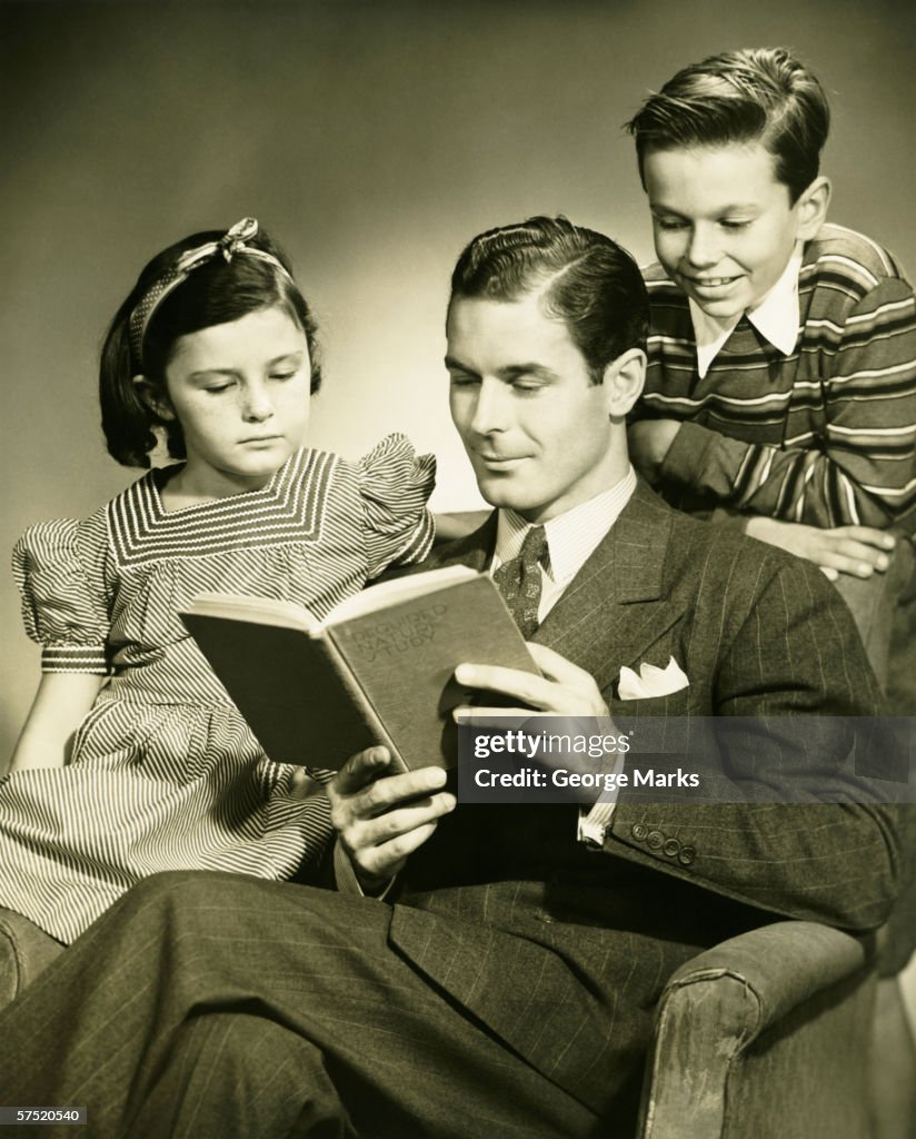 Father reading to son (4-5) and daughter ( 6-7) in studio, (B&W), portrait