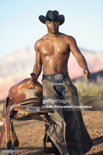 young man in a cowboy hat holding a saddle - calça de couro de cowboy imagens e fotografias de stock