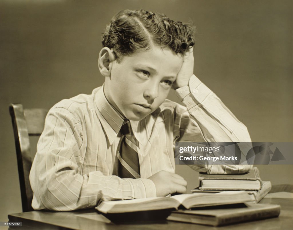 Boy (10-11) sitting at table over open book, head resting on hand, (B&W), close-up