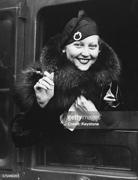 Actress Thelma Todd smoking a cigarette as she smiles from the door of her train carriage as she prepares to leave for America, at Waterloo Station,...