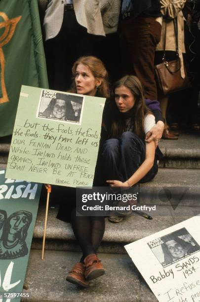 Woman sits on the steps and embraces her daughter while holding a sign featuring the likeness of Bobby Sands and a quote from Padraig 'Patrick'...