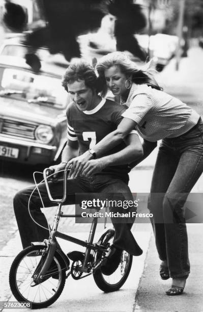 British motorbike champion Barry Sheene tries his hand at a small Chipper bicycle, with the aid of his future wife, glamour model Stephanie McLean,...